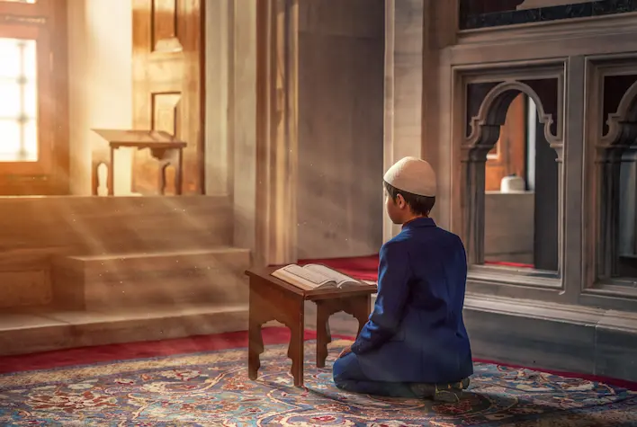 Young Muslim boy sitting on a carpet reading the Quran inside a peaceful mosque to learn Quran online.